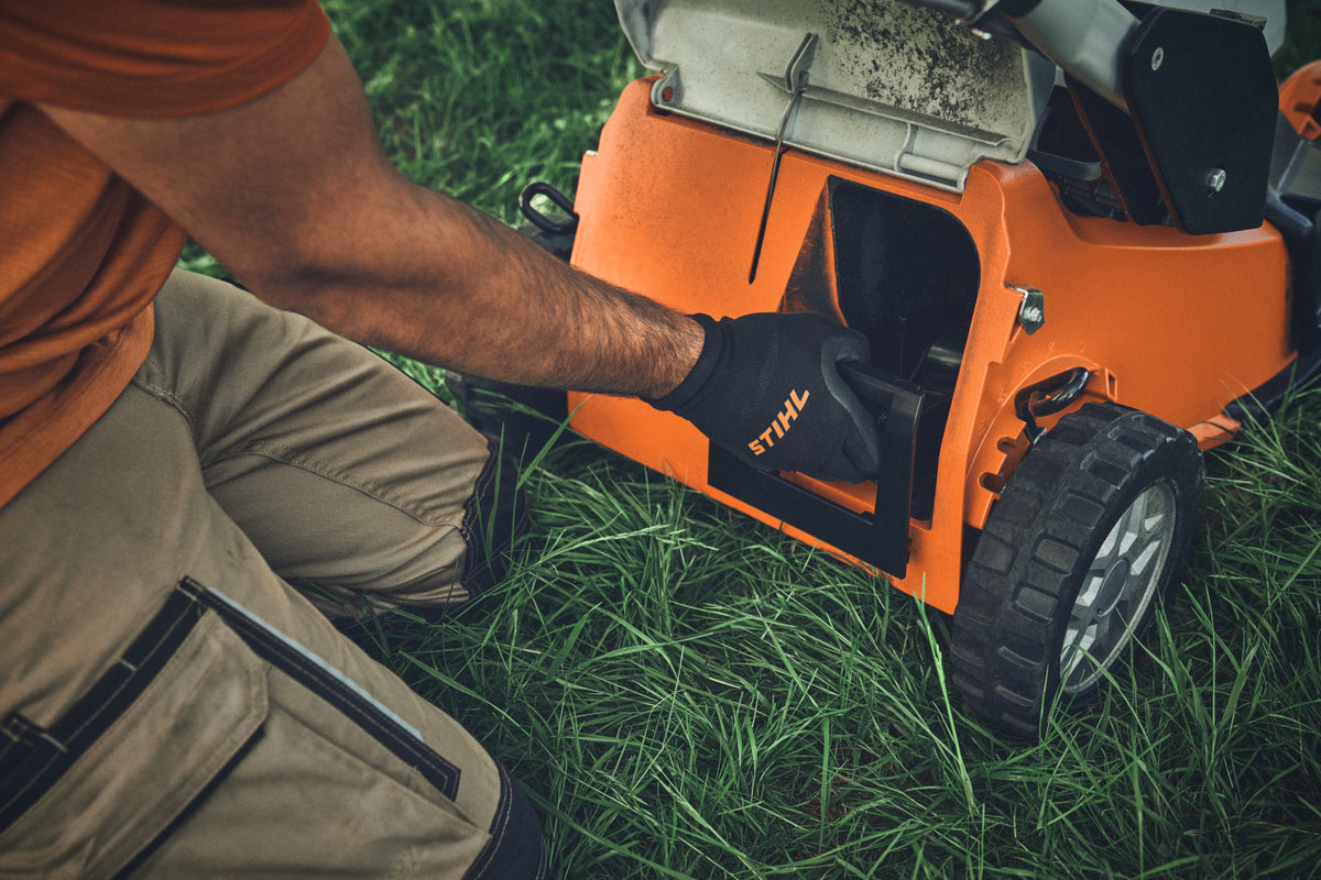 Dettaglio del vano laterale aperto del tosaerba Stihl e della mano guantata dell'operatore.