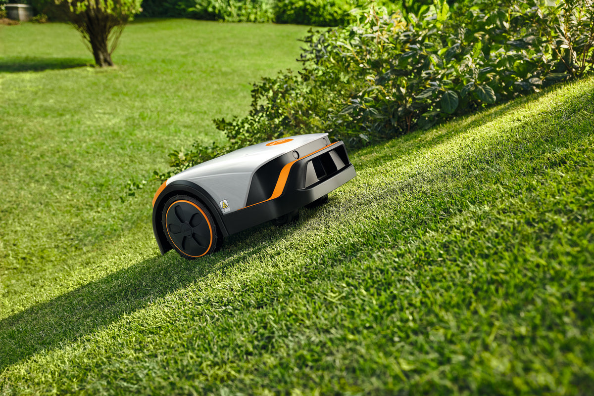 Robot tosaerba Stihl iMOW 4 su un prato in pendenza accanto a una siepe.