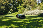 Robot tosaerba Stihl iMOW 4 EVO su un prato in giardino tra alberi e arbusti.